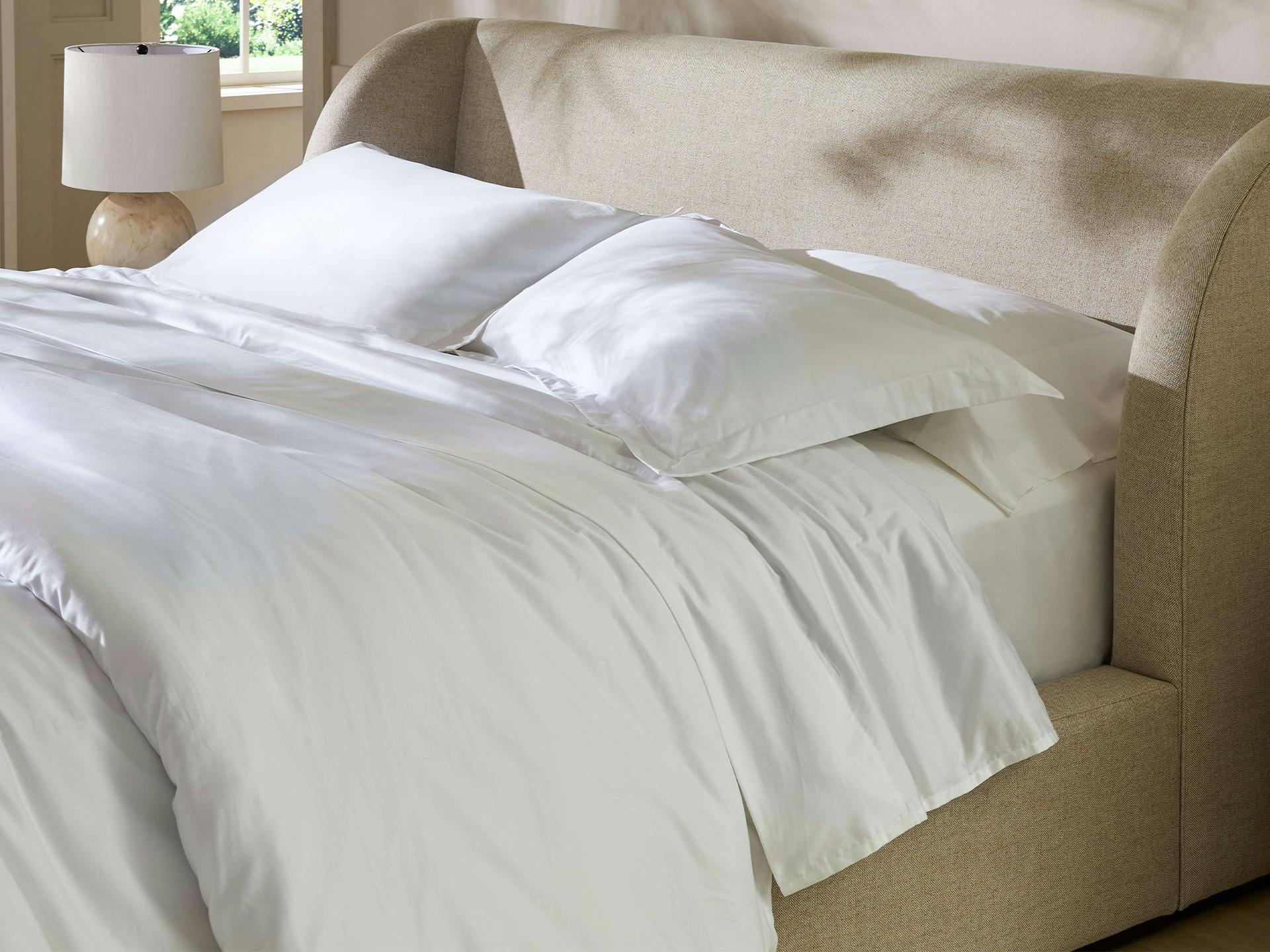 Bedroom scene image brief showing Saatva Organic Sateen Sheet Set on a queen bed in a warm-climate bedroom with fan airflow and thin duvet layer